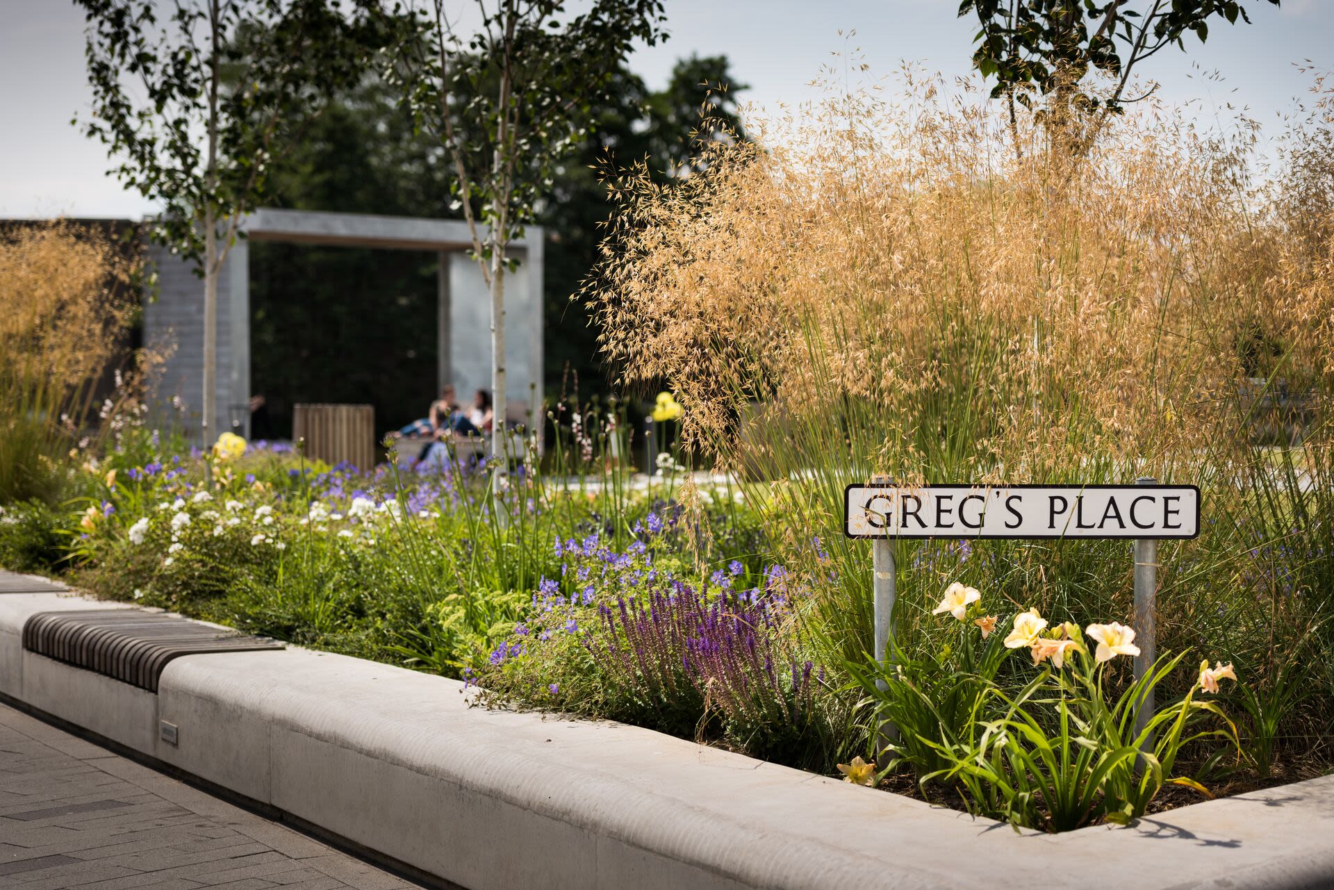 Greg's place on Campus West. Flowers are in bloom next to the sign 'Greg's Place' and two people are sat underneath a large metal shelter.