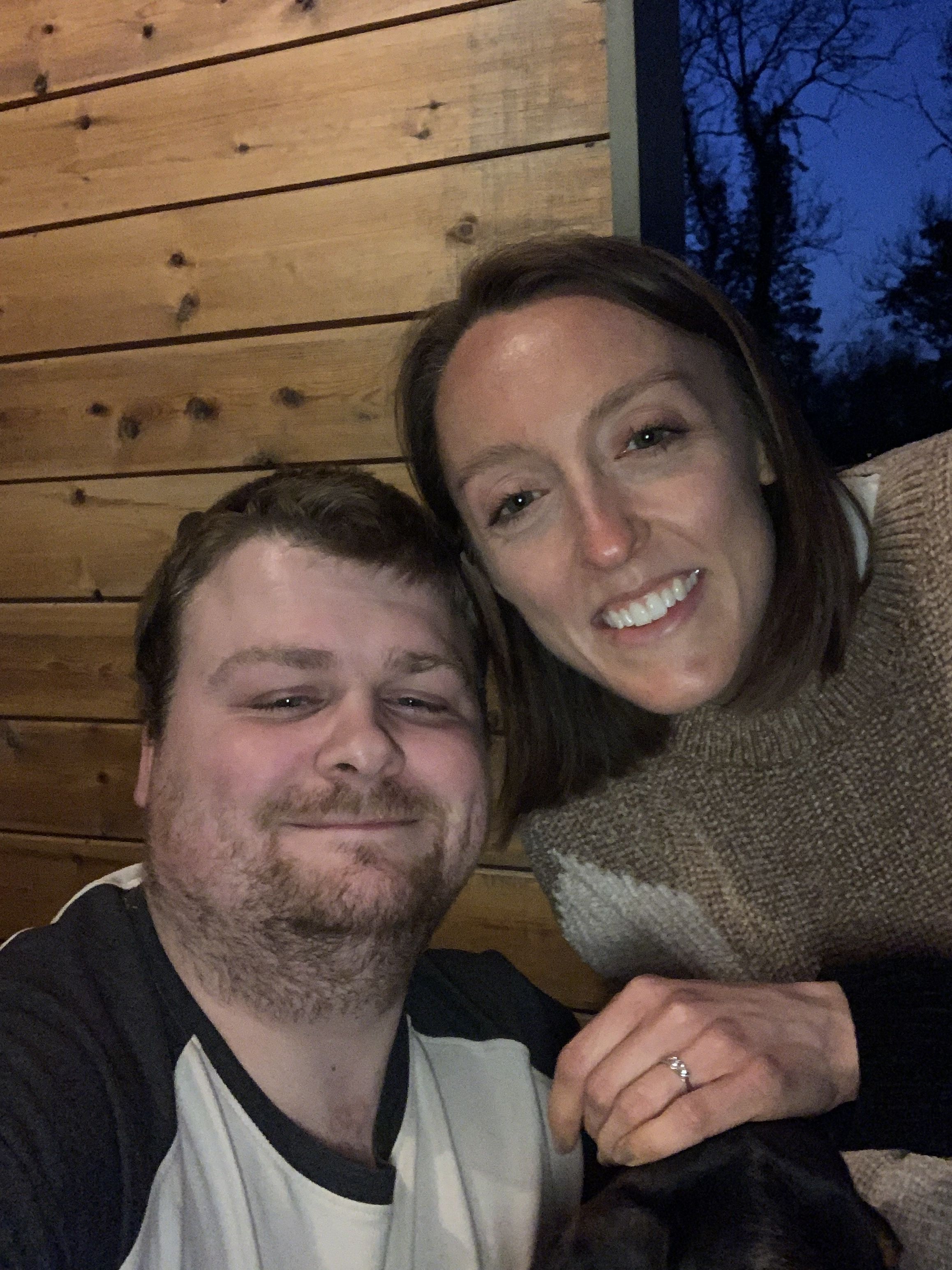 Charlie and Tom pose for a selfie in front of a wood panelled wall. On Charlie's hand is a sparkling engagement ring.