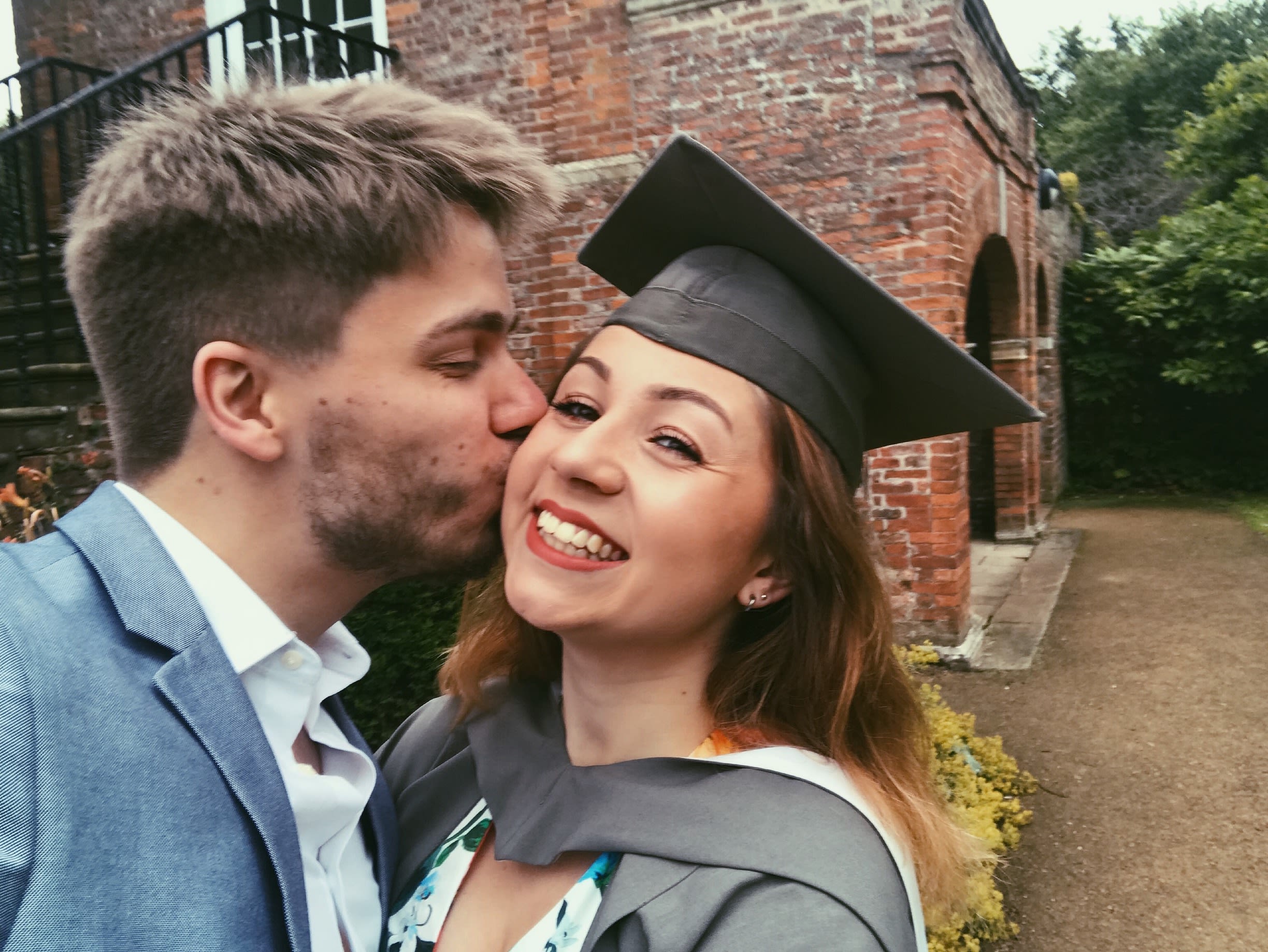 Ashleigh in a York graduation cap and gown outside The Quiet Place. She and Mark are posing for a selfie, and he is kissing her on the cheek.