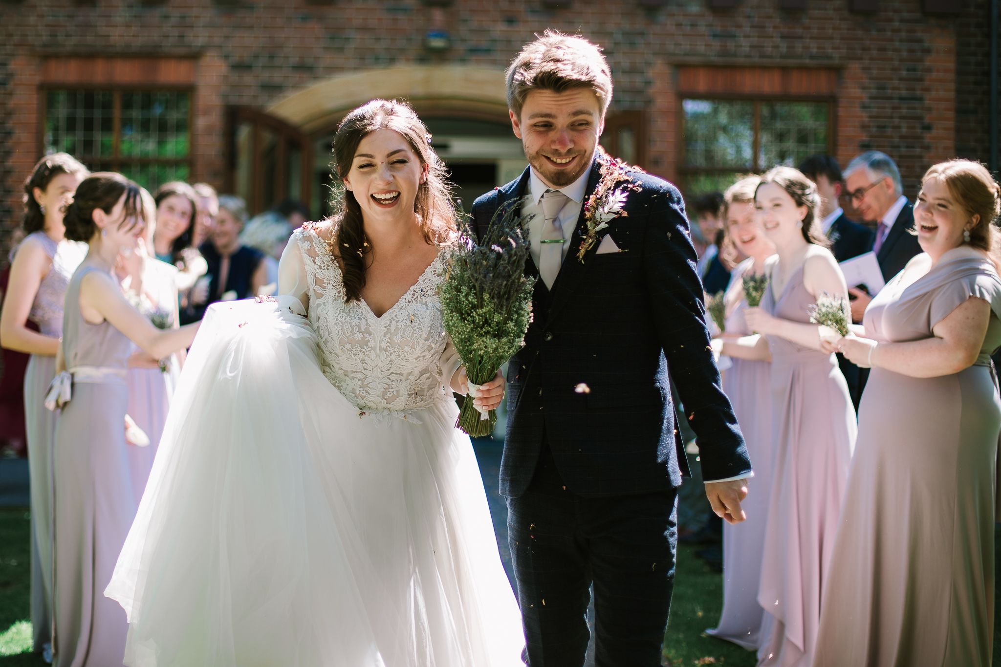 Ashleigh and Mark in wedding attire leaving a red-brick building. Behind them the wedding party cheer.