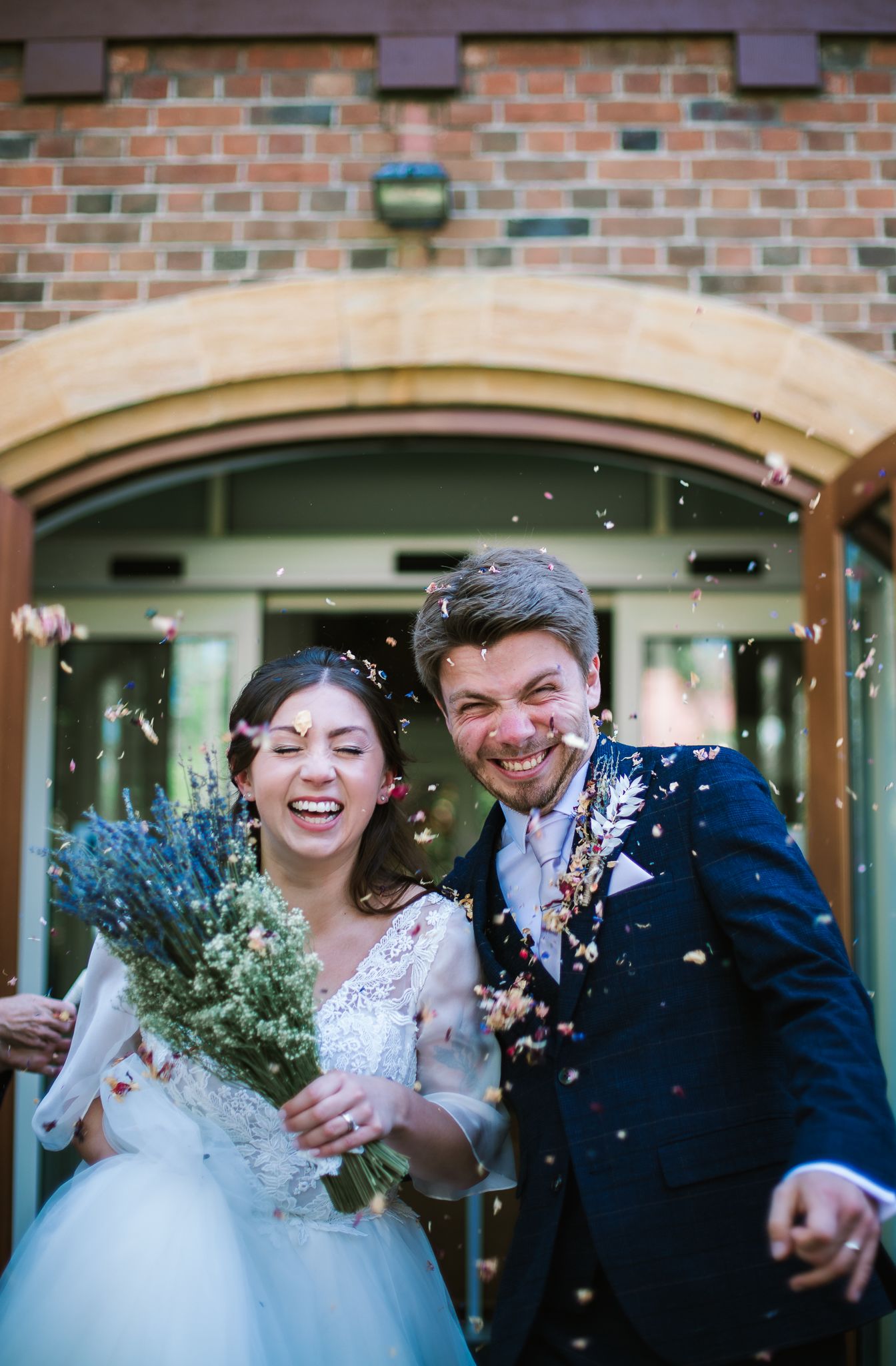 Ashleigh and Mark smile widely as they exit a building on their wedding day. Confetti falls all around them.