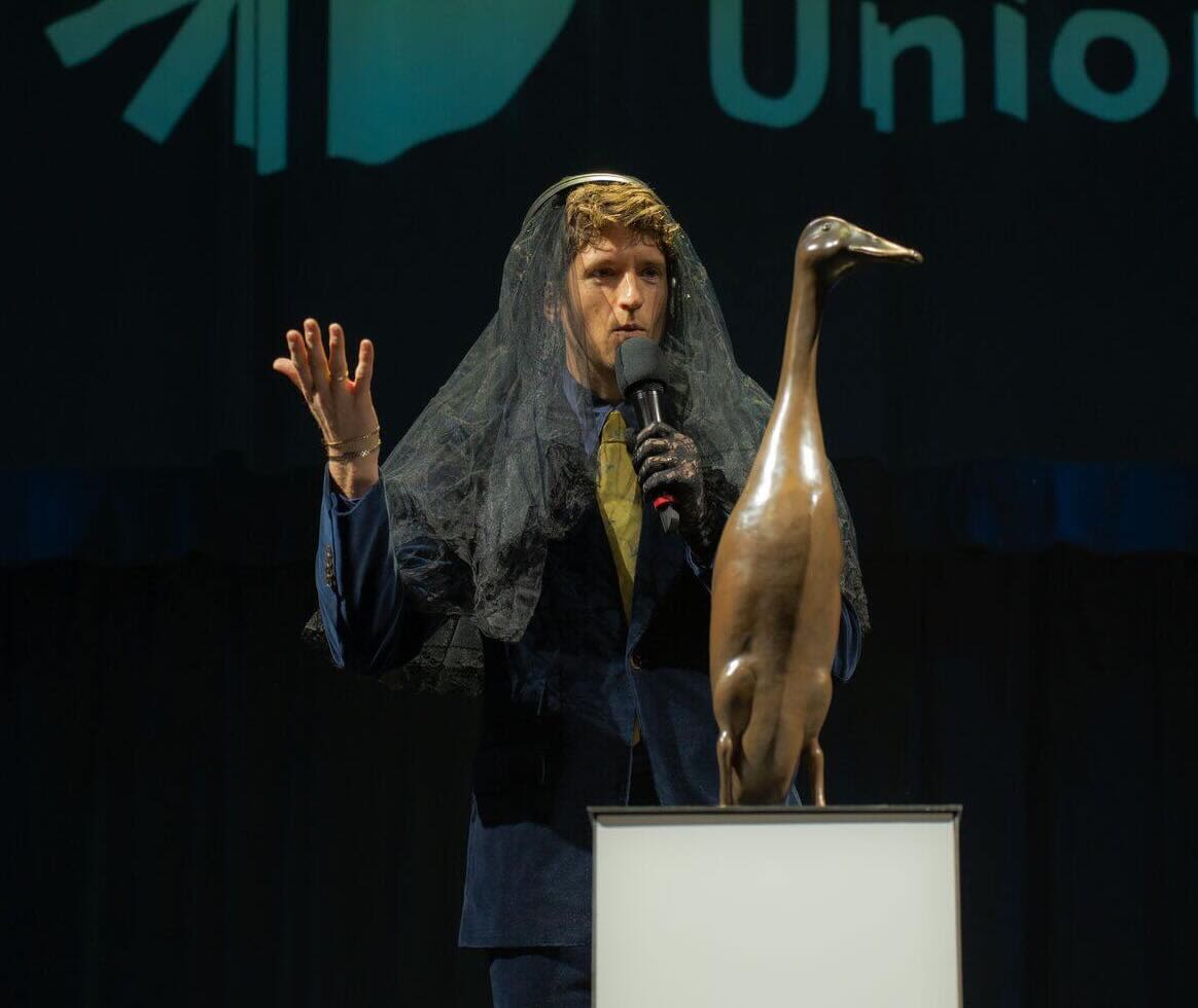 Greg James is wearing a veil and talking into a mic in front of the bronze Long Boi statue