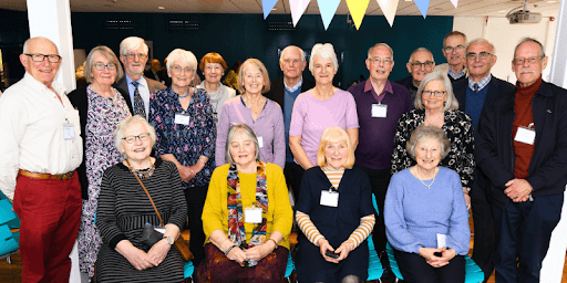 A group of older men and women all stood together in formal clothing, inside Derwent dining room